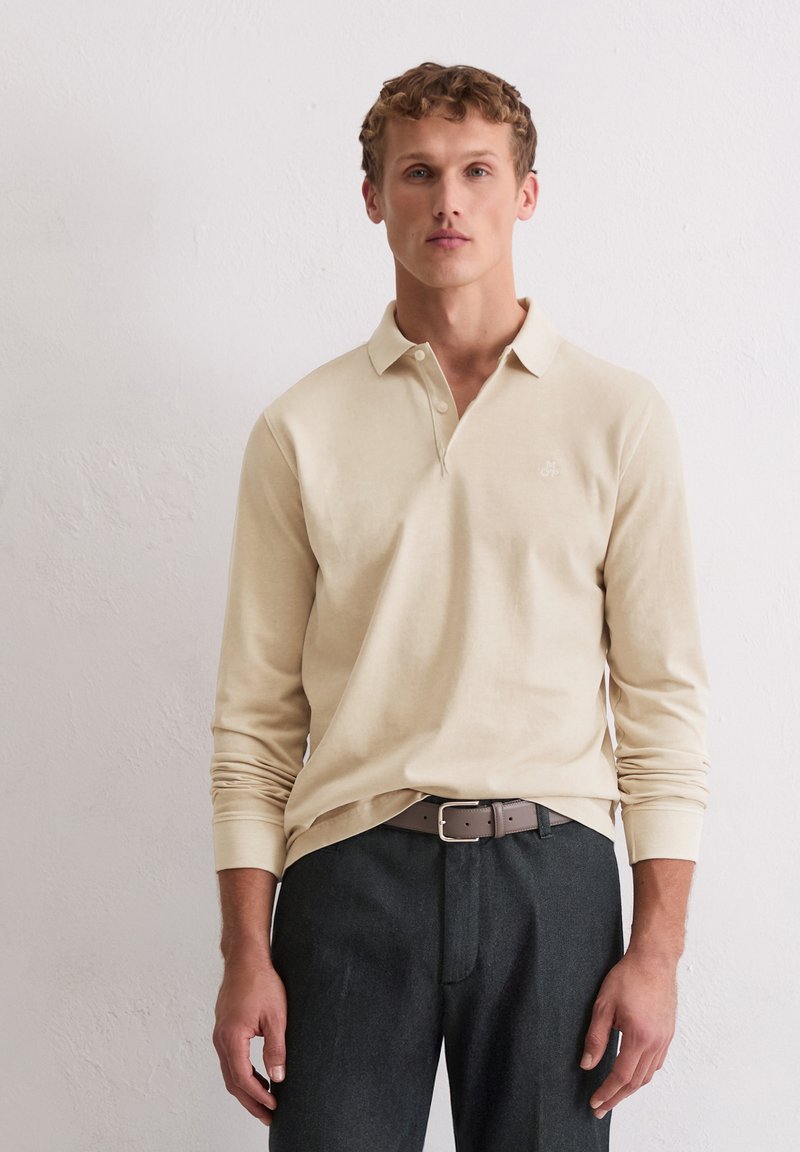 Beige Langarm-Polohemd mit Kragen und einer drei-Knopf-Leiste, kombiniert mit dunkelgrauen Hosen und einem braunen Gürtel. Subtiles Logodesign auf der Brust.