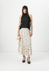 Top sans manches plissé noir, jupe crème avec un motif script noir, escarpins noirs, tenant un petit sac à main beige. Design minimaliste.