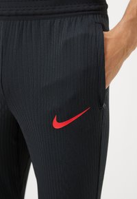Sorte træningsbukser med struktureret stof, der har et rødt Nike-logo på siden og en sidelomme.
