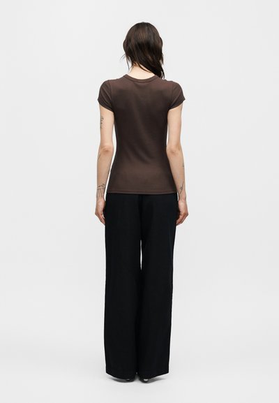 Γυναίκα με μακριά σκούρα μαλλιά, φοράει στενό καφέ κοντομάνικο πουκάμισο και φαρδιά μαύρη παντελόνα, στέκεται με την πλάτη γυρισμένη σε ένα απλό φόντο.