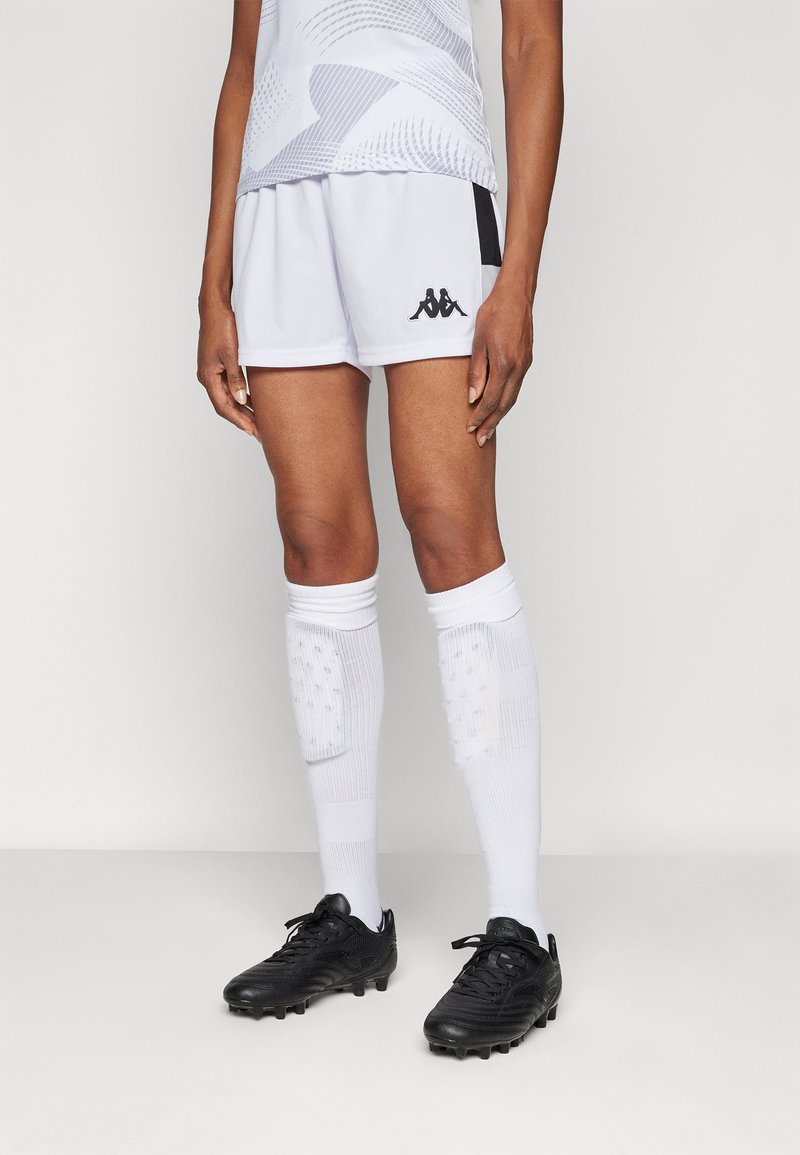 Baltie sporta šorti ar melnu Kappa logo, kombinēti ar melnām futbola kurpēm un baltām ceļgala augstuma zeķēm ar vienkāršām apdrukām.