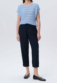 Femme portant un haut en tricot à manches courtes bleu clair, un pantalon court noir à cordon de serrage et des chaussures noires en cuir à enfiler, se tenant sur un fond blanc.