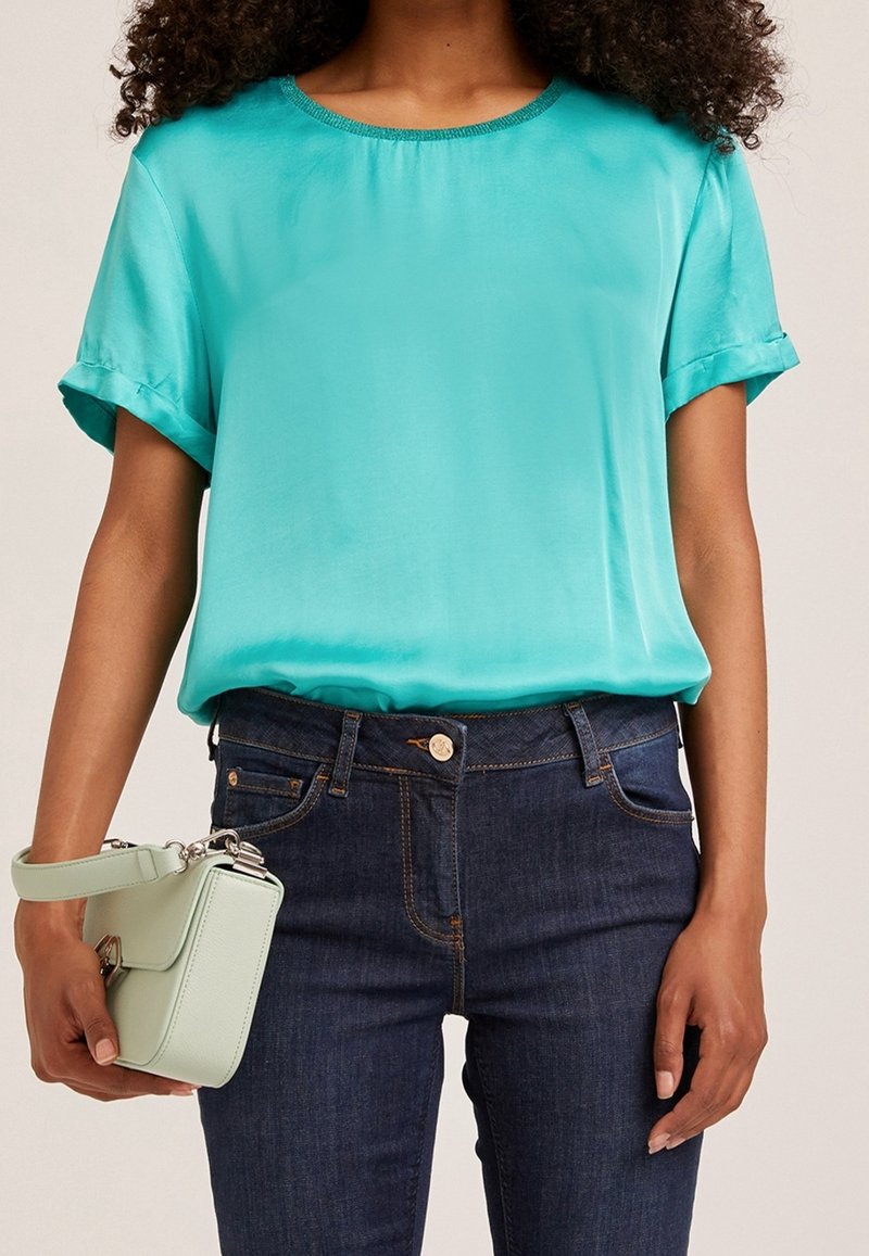 Turkos satinblus med uppvikt ärm, rundad halsringning, parat med mörkblå jeans och en ljusgrön crossbody-väska.