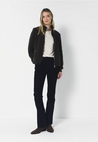 Kobieta stoi ubrana w ciemną rozpinaną kurtkę, białą bluzkę, czarne rozkloszowane spodnie i brązowe buty na jednolitym jasnym tle.