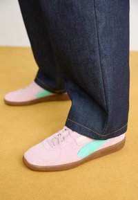 Baskets en daim rose avec un accent vert menthe et une semelle en caoutchouc marron, assorties à un pantalon en denim bleu foncé, sur un fond jaune.