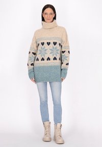 Pulover tricotat bej și teal cu guler înalt, având modele cu fulgi de zăpadă și un design în blocuri de culoare. Asortat cu blugi albaștri deschiși și cizme bej.