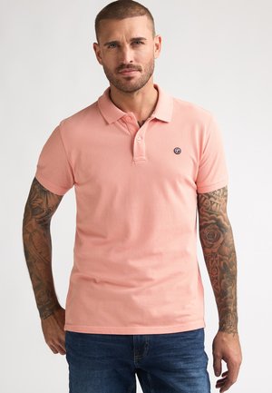 Polo de color rosa claro con una textura suave, que cuenta con cuello, tapeta de dos botones y un pequeño logo en el lado izquierdo del pecho. Combinado con vaqueros azules.