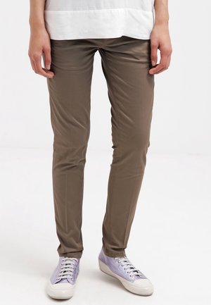 Brązowe spodnie o dopasowanym kroju noszone przez osobę stojącą, zestawione z białymi i jasnofioletowymi sneakersami na sznurowadła oraz białą bluzką.