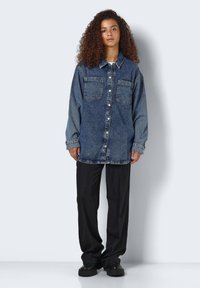 Camicia di denim blu scuro con due tasche sul petto e chiusura con bottoni. Abbinata a pantaloni larghi neri e scarpe nere robuste.