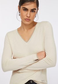 Maglione color beige chiaro con scollo a V in tessuto lavorato a maglia morbido, con bordo a contrasto su scollo e polsini. Presenta maniche lunghe e un dettaglio logo discreto.
