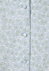 Camicetta a maniche lunghe in stoffa azzurra chiara con un motivo floreale di fiori bianchi e foglie verdi, dotata di chiusura centrale con bottoni.