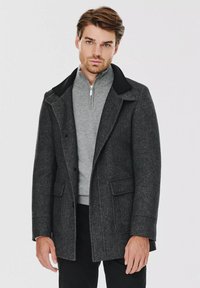 Szary płaszcz z wełny z czarnym kołnierzem, zapinany na podwójne guziki, z dwiema kieszeniami z przodu. Wewnątrz widoczny jest jasnoszary sweter na zamek.