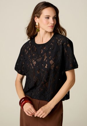 Femme portant un chemisier en dentelle noire à motif floral, un pantalon marron, de grands bracelets rouges et des boucles d'oreilles dorées, regardant légèrement de côté sur un fond uni.