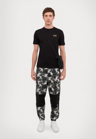 T-shirt en coton noir avec logo jaune, associé à un pantalon camouflage aux motifs verts et blancs, et des baskets blanches. Sac en bandoulière inclus.