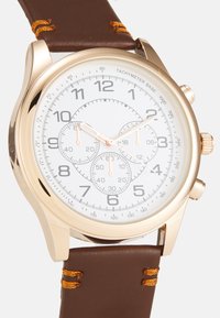 Reloj dorado con una esfera blanca. Cuenta con tres subesferas, números oscuros y una correa de cuero marrón con detalles dorados cosidos.