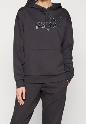 Person iført mørkegrå Puma-hættetrøje og matchende bukser med zebra-stribet Puma-logo på brystet og hænderne i lommerne.