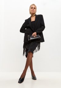 Blazer nero su un vestito nero senza spalline con dettagli a frange; abbinato a tacchi neri e una clutch nera, che mette in mostra una silhouette elegante.