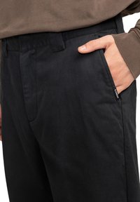 Čierne bavlnené nohavice s uvoľneným strihom, prednými vreckami a diskrétnym logom. Materiál má hladkú textúru v klasickom dizajne.