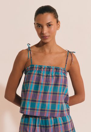 Femme aux cheveux attachés, portant un top sans manches à carreaux bleu et violet avec de fines bretelles et un bas assorti, debout devant un fond uni.