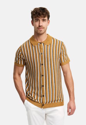 Homme portant une chemise à manches courtes jaune moutarde à rayures verticales noires et blanches et un pantalon blanc, debout avec une main dans la poche.