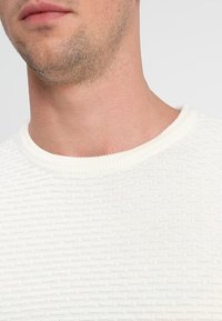 Lightweight, textured white sweater with a ribbed crew neckline. Features horizontal patterns and soft fabric. Suitable for casual wear.