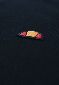 Un tissu noir présente un petit logo brodé avec un design en demi-cercle en orange et rouge, posé sur un fond texturé.