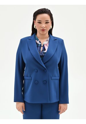 Donna che indossa un blazer doppiopetto blu con pantaloni abbinati, una camicetta floreale sotto e orecchini dorati vistosi, rivolta verso l'obiettivo.