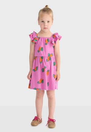 Ragazza giovane in piedi che indossa un vestito rosa con stampe di carote e scarpe color cuoio con lacci rosa acceso su uno sfondo chiaro e semplice.