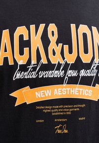 Črna bombažna majica s kratkimi rokavi z velikim oranžnim napisom in belimi poudarki. Dizajn vključuje stilizirano ime blagovne znamke in grafične elemente.