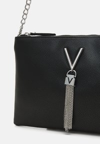 Černá kožená psaníčko s texturou a stříbrným řetízkem, které má výrazný stříbrný akcent ve tvaru "V" a detail řetízku níže. Zavírání na zip na vrchu.