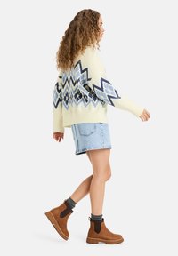 Kremgenser med blå geometriske mønstre, lyse denimshorts og brune ankelstøvler med ribbede detaljer. Krøllete hår stylet ned.
