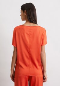 Red short-sleeve top in smooth fabric, featuring a wide neckline and an uninterrupted back seam, worn with coordinated wide pants.