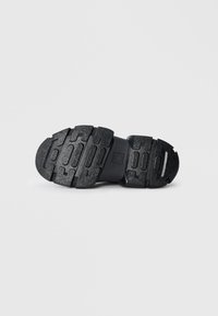 Semelle de chaussure en caoutchouc noir avec surface texturée, présentant des rainures multidirectionnelles et un design segmenté pour une meilleure adhérence.