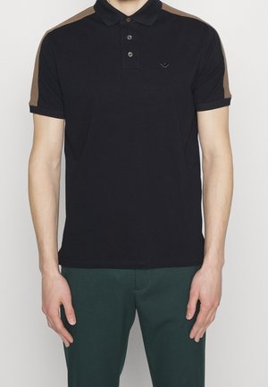 Mężczyzna w czarnej koszulce polo z brązowymi paskami na ramionach i ciemnozielonych spodniach, stojący z rękami swobodnie opuszczonymi wzdłuż ciała.