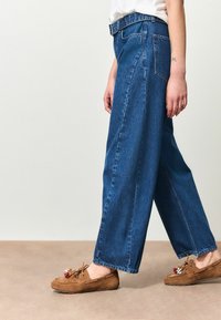 Blauwe wijde spijkerbroek met een hoge taille, voorzien van het klassieke ontwerp met vijf zakken en subtiele stiksel details. Gecombineerd met bruine mocassins.