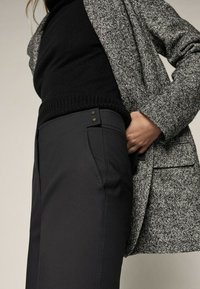 Grijs wollen blended jas over een zwart gebreid sweater, gecombineerd met hoge zwarte broeken met contrasterend stiksel en metalen accentdetails.