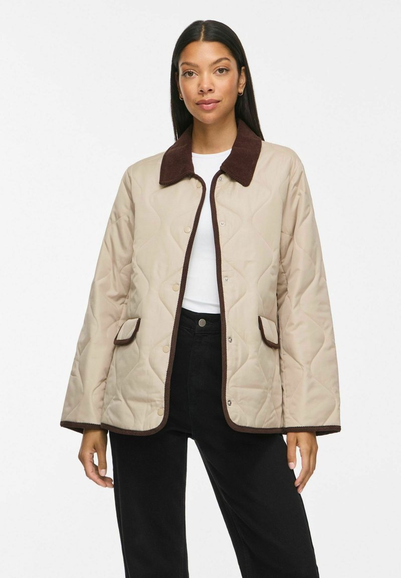 Femme portant une veste matelassée beige avec col, rabats de poche et bordure marron foncé sur chemise blanche et pantalon noir sur fond blanc.