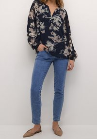 Nainen, joka on pukeutunut läpinäkyvään tummansiniseen kukkablousiin, vaaleansinisiin kapeisiin farkkuihin ja beigeihin kärjen avokorkoihin, seisoo yksinkertaista valkoista taustaa vasten.