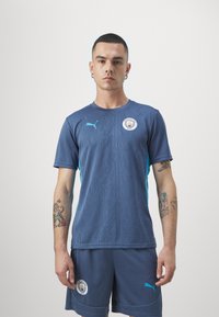 Zila sporta krekls ar teksturētu rakstu un tirkīza akcentiem, uz kura ir Mančestras City logo. Saskaņots ar atbilstošām biksēm.
