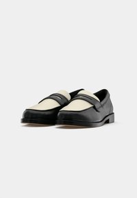 Par de mocassins em couro preto e bege com salto baixo e detalhe de tira tipo penny, exibidos sobre um fundo branco.