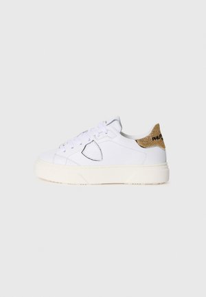 Baskets blanches avec un bout arrondi, des lacets, et un accent doré texturé au talon. Elles présentent une semelle en caoutchouc et des détails de couture subtils.