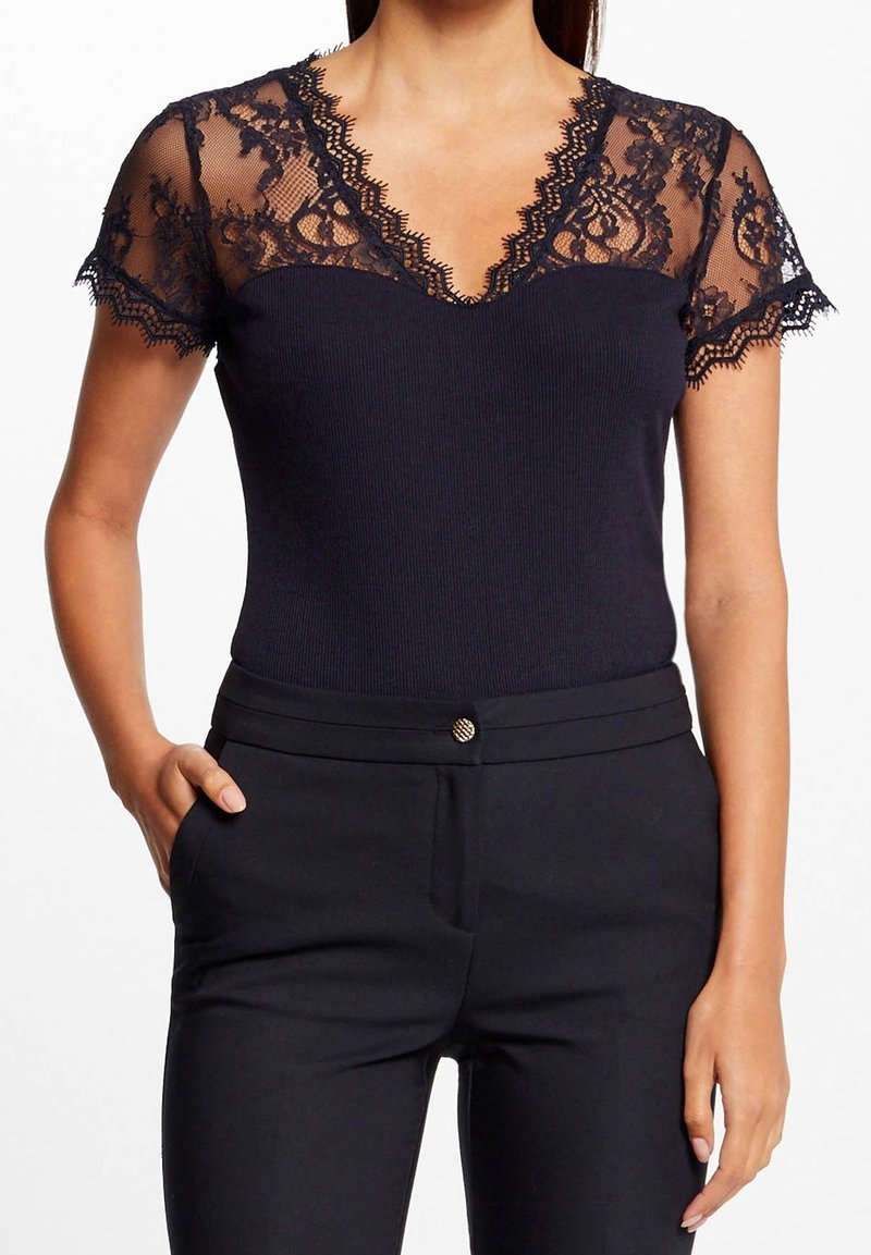 Femme portant un haut noir moulant avec des manches courtes en dentelle florale transparente et un pantalon noir taille haute assorti avec détail de boutons.