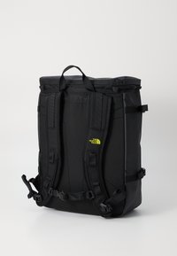 Sac à dos noir avec des bretelles rembourrées, une poignée sur le dessus et des boucles sur les côtés. Comprend un matériau texturé et un petit logo jaune à l'avant.