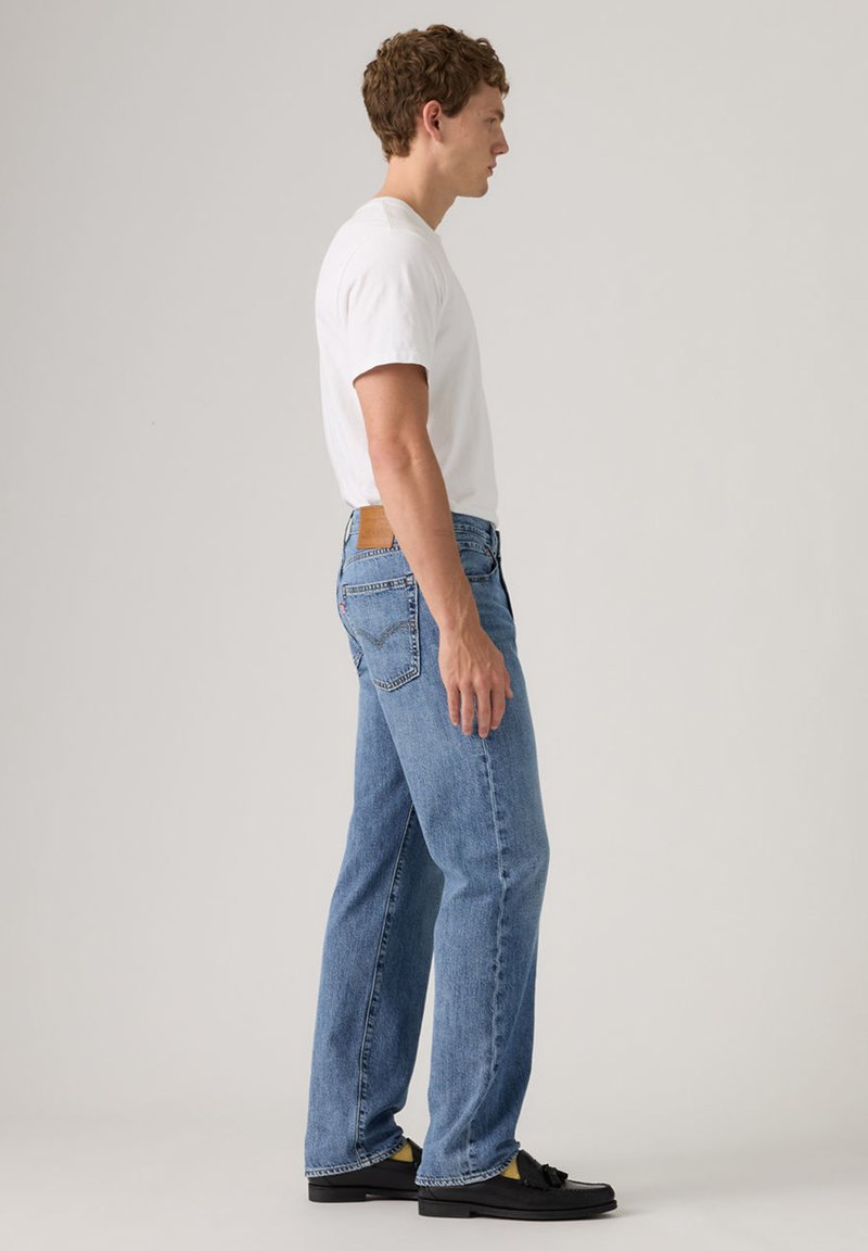 Homme debout de profil portant un t-shirt blanc, un jean bleu et des mocassins noirs sur un fond clair uni.