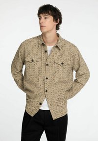 Koszula w odcieniu brązowym z wzorem, o teksturowanej powierzchni, z zapinanym przodem, dwiema kieszeniami na piersi i czarnymi guzikami, noszona na białej koszulce.