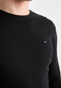 Närbild av en person som bär en svart långärmad skjorta med en liten röd, vit och blå logotyp på vänster bröst.