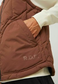 Brown quilted vest with a side pocket and R.D.D. text detail, featuring sleek stitching and a soft, padded texture.