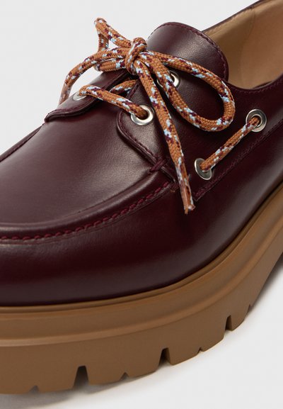 Bordeaux læder loafers med tyk tan gummisål og brune mønstrede snørebånd bundet i en sløjfe gennem sølvøjer.
