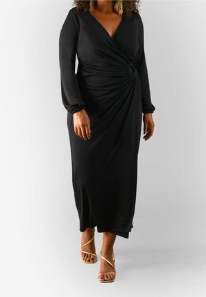 Vrouw draagt een lange zwarte wikkeljurk met geplooide zijkant en gouden sandaaltjes met hoge hak en bandjes, lopend tegen een effen achtergrond.