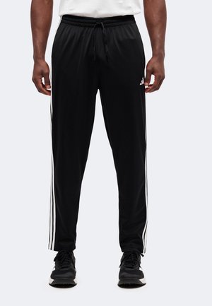 Homme portant un pantalon de survêtement Adidas noir avec des bandes blanches sur les côtés et des baskets noires, debout devant un arrière-plan uni.